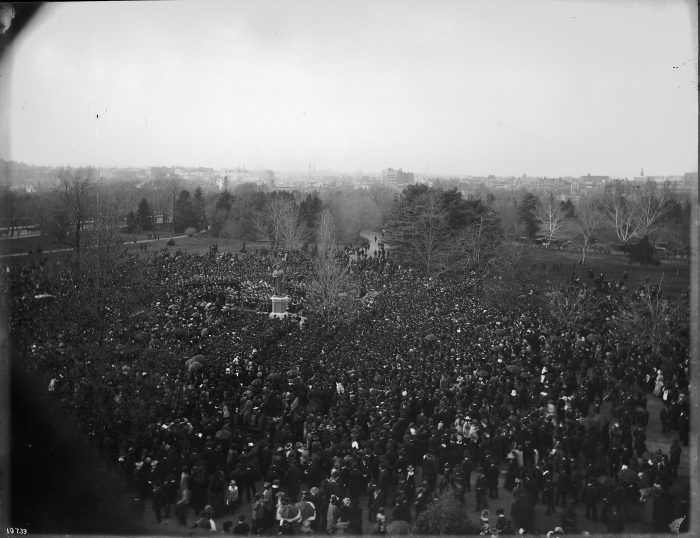 The dedication ceremony of the Joseph Henry statue with a large number of people gathered around the statue on the Mall with the skyline of Washington, D.C., in the background, 1883. The photograph was taken from one of the towers in the Smithsonian Institution Building