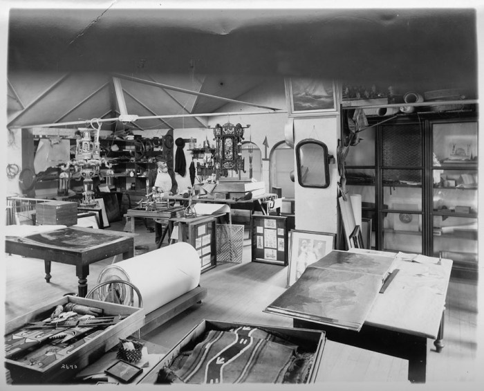 A work and storage area on the third floor of one of the pavilions in the United States National Museum, now the Arts and Industries Building, ca. 1890. Looking to his left, a museum employee stands at a table with an object he is working on. Stored in the room are various artifacts, including works of art, a mirror, Native American objects, and Asian lanterns.