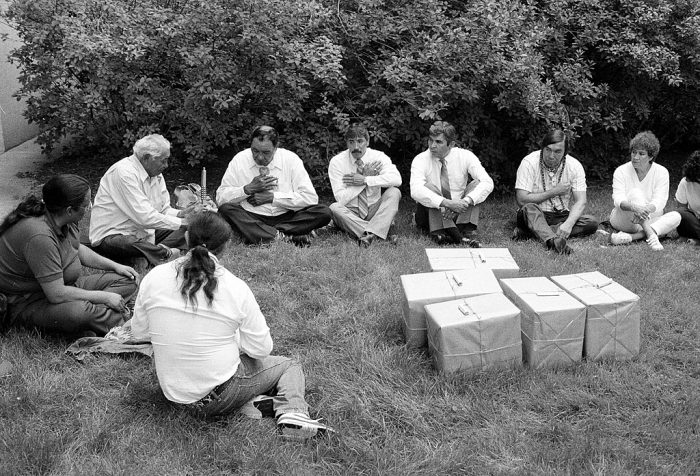 Group passes pipe while sitting on ground near several boxes of repatriated remains