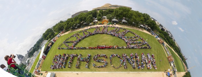 Staff of Smithsonian Institution pose spelling out SMITHSONIAN with a half-sunburst pattern at the 2015 Smithsonian Folklife Festival, Washington, D.C., June 30, 2015. Composite panoramic photograph with full sky area by Smithsonian National Air and Space Museum Photographer Dane Penland. [SmithsonianStaffPhoto6-30-2015-Pan5 wide-2BEST-FullSky] [NASM2015-04136]