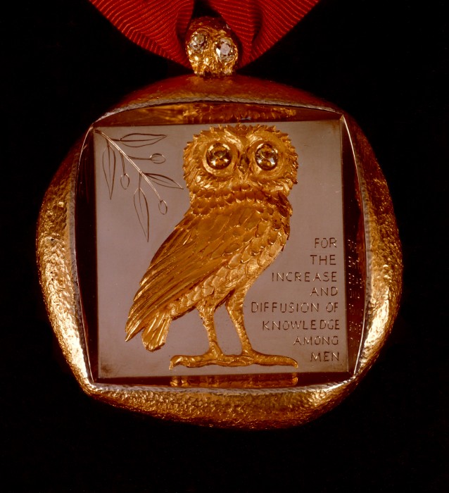The Smithsonian’s Badge of Office is a thick, irregularly shaped medallion made of 18-carat gold that hangs from a cherry-red ribbon. Cast in high relief and set within a deeply recessed square on the front is the owl of Athena, a symbol of wisdom, and an olive branch, a symbol of peace and goodwill. The owl has forward-gazing eyes that are set with a pair of large, yellow sapphires. To the side of the owl is engraved the Smithsonian Institution’s mandate as defined in James Smithson’s will: “For the Increase and Diffusion of Knowledge Among Men.” The back is engraved with the sunburst seal of the Smithsonian and “James Smithson, 1765—Bicentennial 1965.” (Image courtesy Smithsonian Institution)