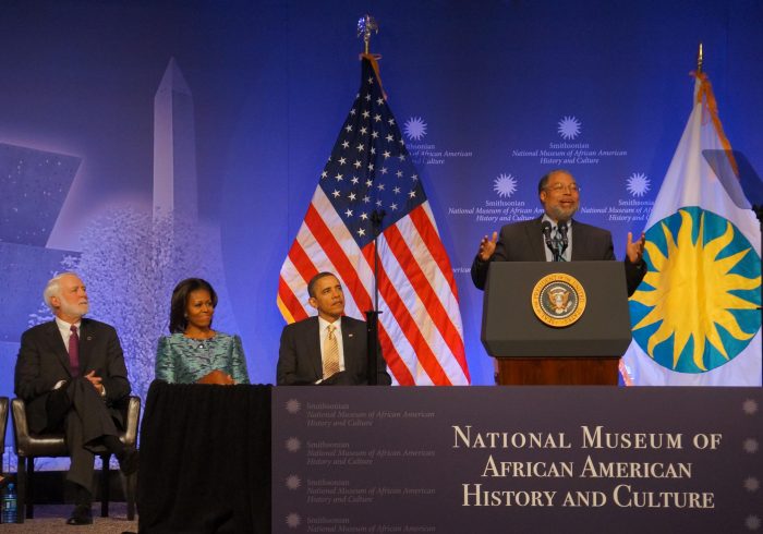 Bunch at podium with US flag and dignatiries behind him