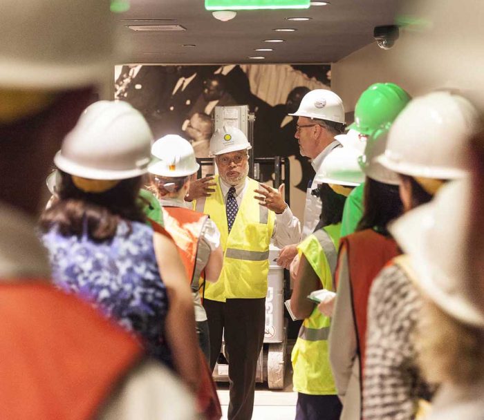 Bunch, in hard hat, facing camera while others with backs to camera listen