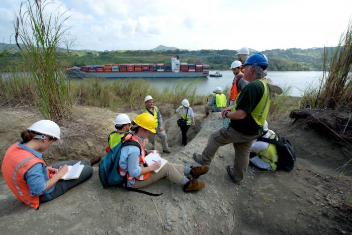 People in hard hats and vests take notes on the bank of the Panama Canal with a cargo ship in the background