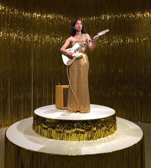 WOman in gold dress holds a guitar atop adais covered with gold foil