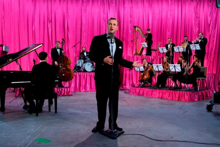 Still image showing artist at mic with orchestra and hot pink curtain behind him