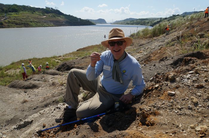 Skorton, wearing hat and sunglasses, smiles into camera and holds up a small fossil. Panama Canal in background