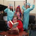 Poster sits at desk as friends in Statue of Liberty costumes pose behind her