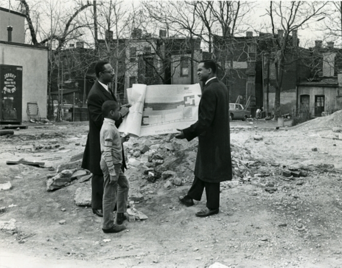 Two men and child look at blueprints in vacant lot