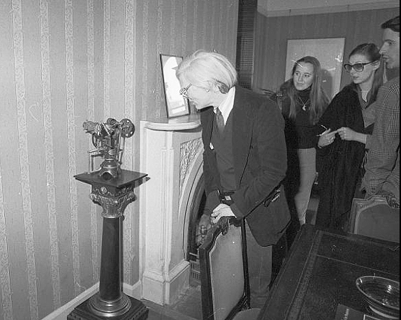 Warhol looking at elaborate model on pedestal. Unidentified women in background