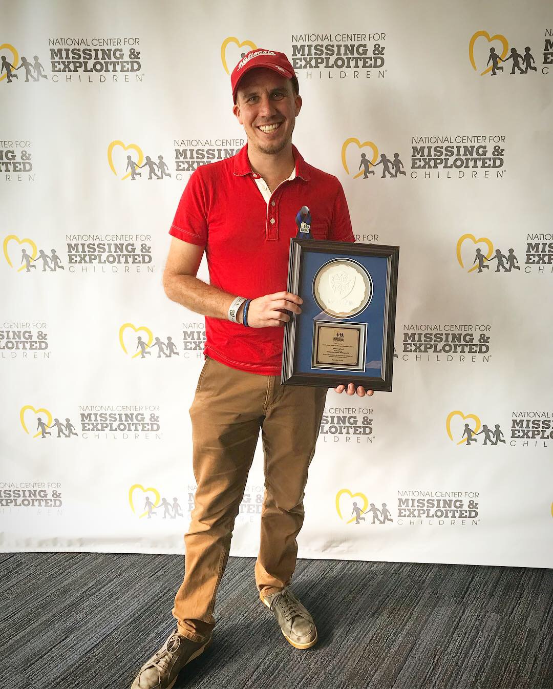 Gagliardi in red shirt and cap in holding award in front of step-and-repeat screen