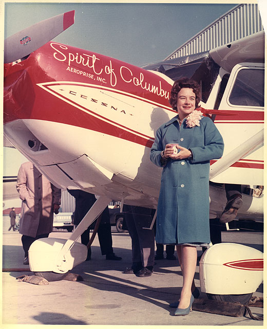 Mock in coat dress and corsage poses beside her plane