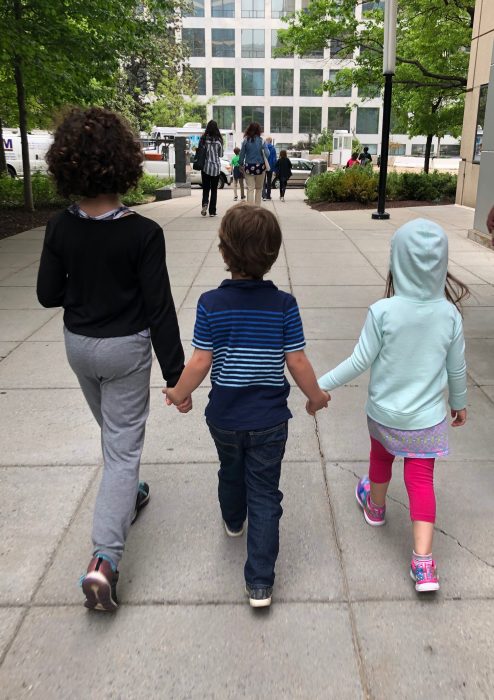 Three young children walk hand-in-hand away from the camera