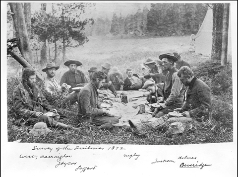 Black and white photographic print of a group of individuals seated outside, on the ground, at mealtime. Trees and a tent can be seen in the background. The group, on the Hayden Survey of the Territories in 1872, includes West, E. Campbell Carrington, Thomas W. Jaycox, Taggart, Clifford De V. Negley, William H. Jackson, William Henry Holmes on the right side, and Beveridge. Their names are handwritten underneath the image.