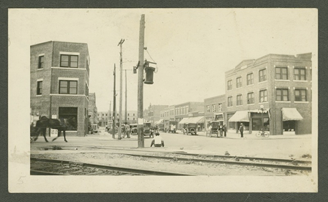 Black-and-white photograph of North Greenwood Ave. in Tulsa, Oklahoma. The photograph was taken looking North down the avenue from East Archer Street. The Williams Building is visible on the left side of the photograph. Across the street the east side of N. Greenwood Ave. is visible. On the far right side of the photograph is the Botkin Building. Several people are seen walking or standing around the street. There are several automobiles parked on along the street. A horse in carriage is on the left side of the photograph. A man dressed in a white shirt is sitting down on the curb of E Archer St. in the center of the photograph.