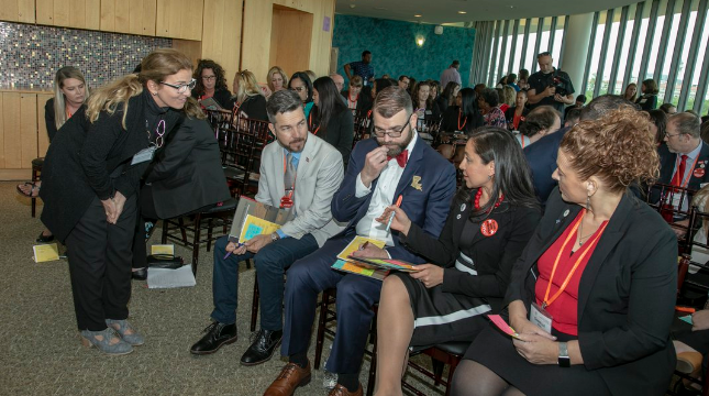 Blonde woman talks to seated men and women at conference