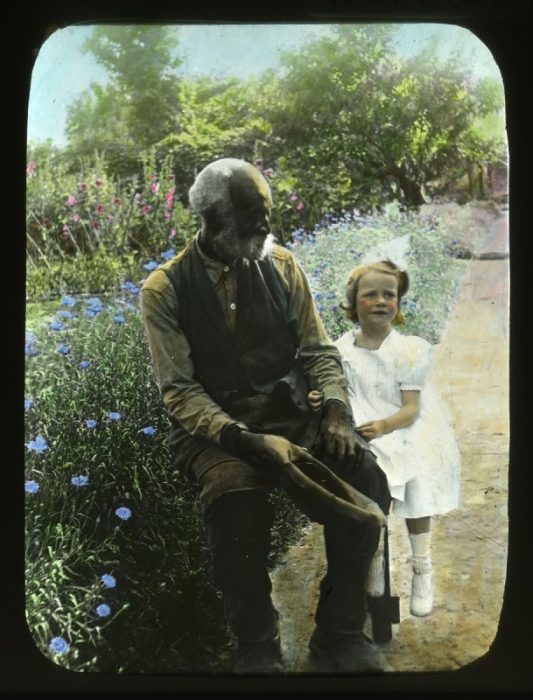 Hand tinted photo of old black man and little white girl in garden