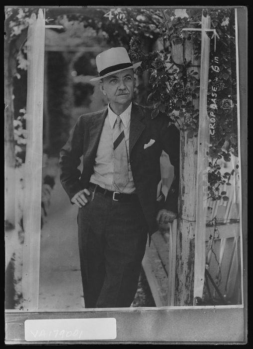 Man in suit and hat poses under rose arbor