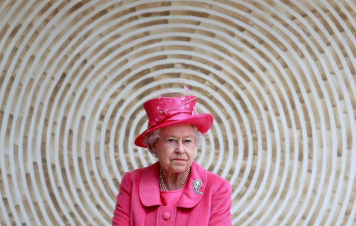 Queen in pink hat and suit with concentric circle architectural detail behind her