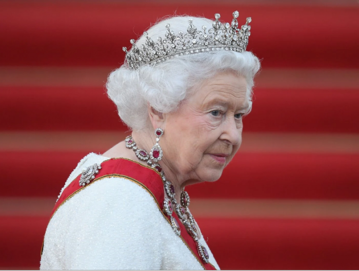 Queen in white wearing crown and jewels against a red background