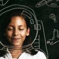 Smiling young girl poses against blackboard featuring chalk drawings of astronauts and space