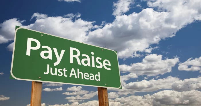 Green traffic sign saying "Pay Raise Just Ahead" against blue sky with white clouds