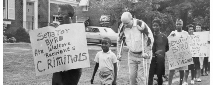 Small group of protesters inclusing white man on crutches young Black boy, led by woman carrying sign