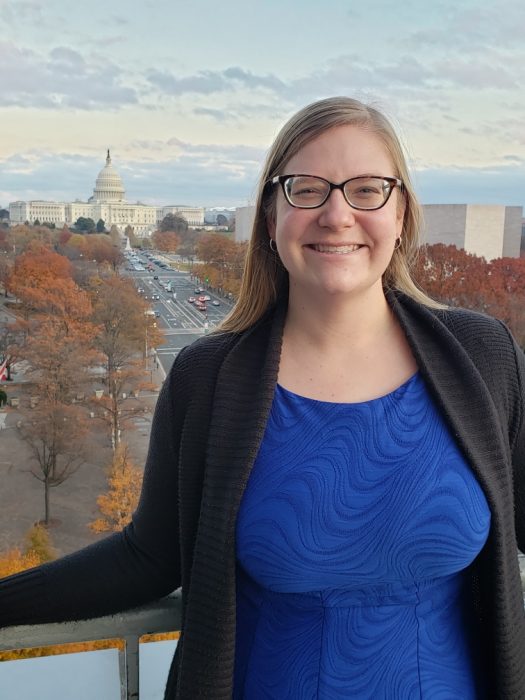 Michelle Gelhausen in blue shirt and sweater poses with US Capitol in background