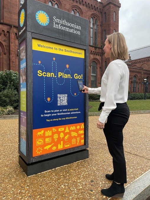 Woman in black shirt and white pans uses her phone to scan code on Visitor Information pylon