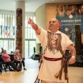 Cherokee man in traditional dress addresses crowd in Potomac Atrium of NMAI
