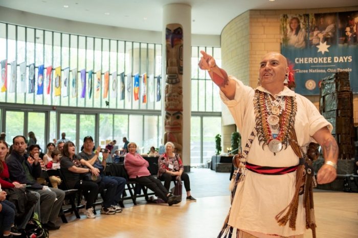Cherokee man in traditional dress addresses crowd in Potomac Atrium of NMAI