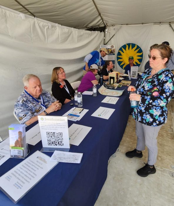 Volunteers behind desk talking to visitors in tent, Smithsonian sunburst logo in background