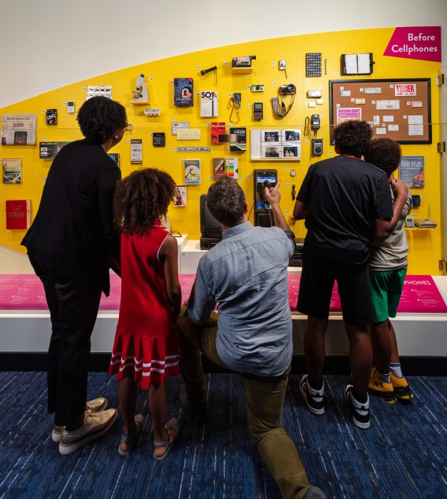 Family listens as kneeling man points to and explains artifacts in "Before Cellphones" display