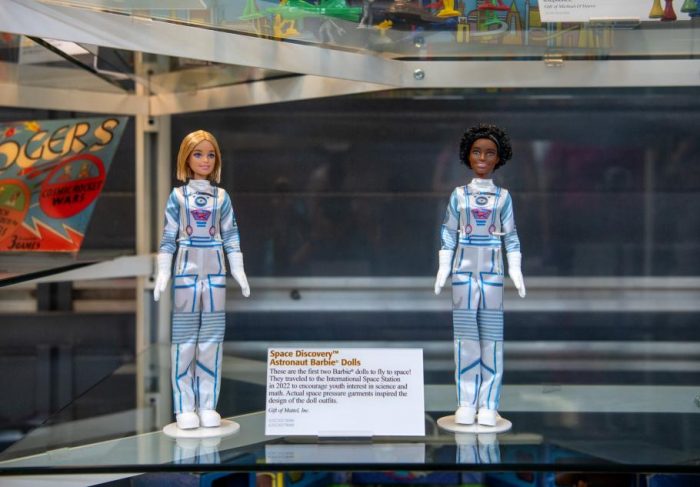 Blonde and African American Barbie dolls in ISS suits on display with "Space Discovery Astronaut Barbie" information card