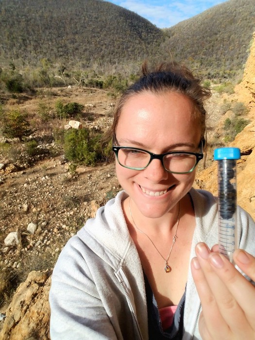 Photo of Sally Bornbusch in an outdoor setting. She is smiling to the viewer and holding a sealed container with animal feces in it.