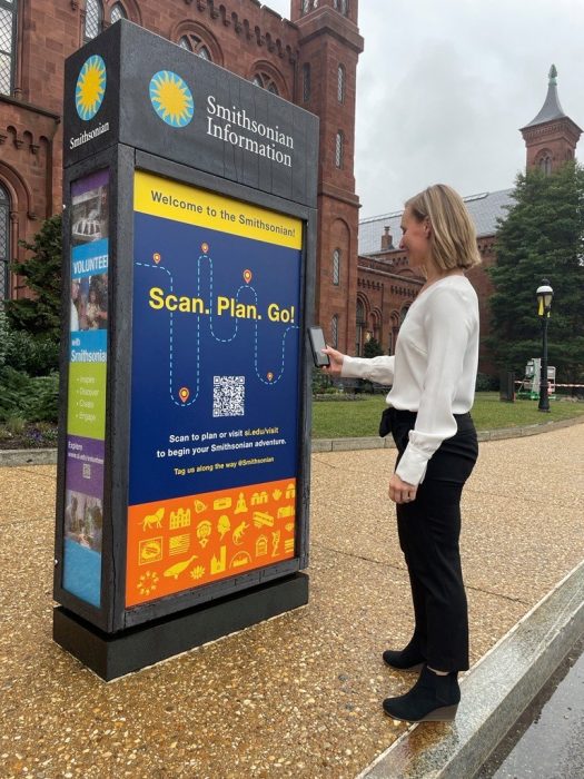 Woman in white shirt and black pants scans a QR code at the Scan, Plan, Go kiosk