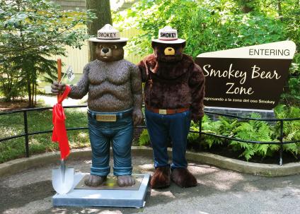 Costumed Smokey Bear stands next to statue of the character next to "Smokey Bear Zone" sign at the National Zoo in 2019.