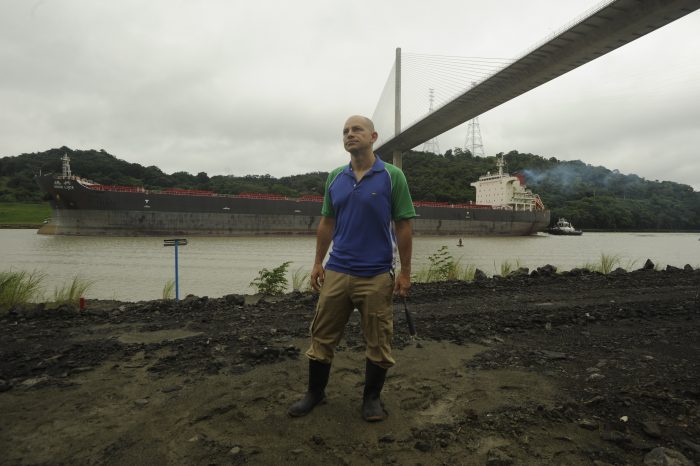 Jaramillo wearing boots on side of Panama Canal as cargo ship passes in the background