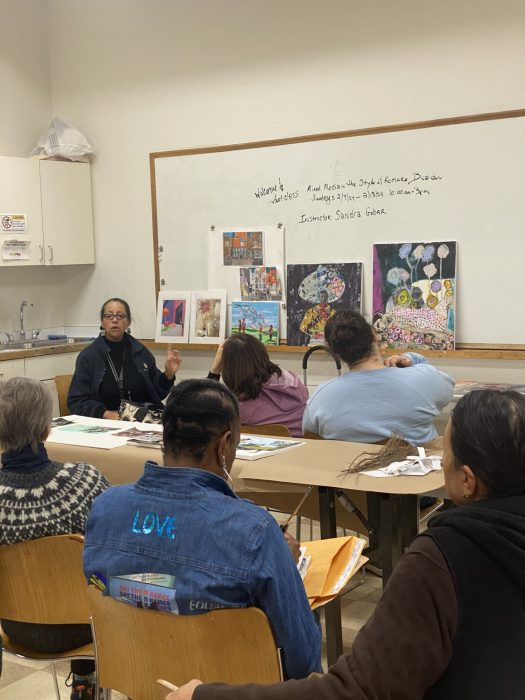 Instructor address adult students; artworks displayed on whiteboard behind her