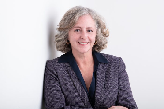 Formal Portrait of Elizabeth Babcock, wearing grey suit, leaning against a white background