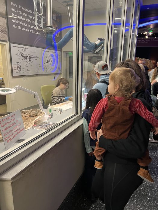 Visitors, including woman holding a toddler watch Myria work in the Fossil Lab