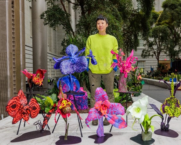 Aretis in brigt green shirt stands behind several of her sculptural models of orchids