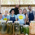 A group of people, some in pale blue Folklife Festival Volunteer T-shirts, pose behind a row of jarred pickled vegetables.