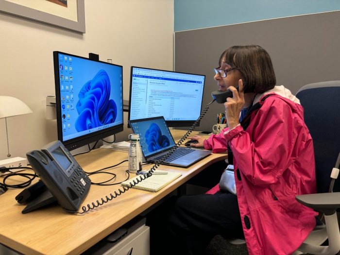 Deloreto, wearing pink jacket, answers the phone while sitting at desk