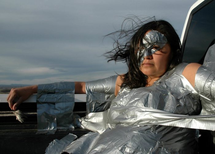 Photo of artist masked and wearing flowing silver garment with overxast sky in the background