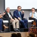 Kirsten Hall, in red shirt and funky shoes, sits on a panel with several older men.