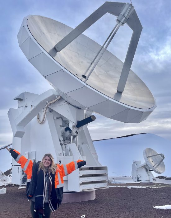 Kirsten hall raises her arms in jubilation in front of the Submillimeter Array Telescope in Hawaii