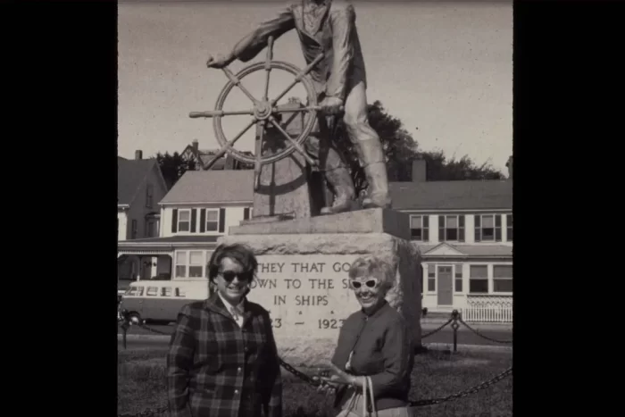 Nlack and white photo of two women wearing sunglasses in front of Fisherman's Memorial statue in Gloucester, Mass.