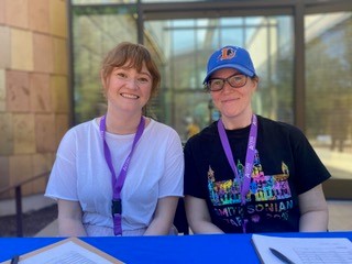 larissa Kunynskyj and Ashley Grady, wearing purple lanyards, smaile for the camera while sitting at the registration desk.