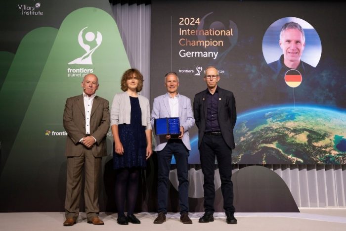 Four people stand on stage, Ellen Welti and Peter Haas are flanked by two unidentified men. Background scrims read 2024 International Champion Germany with a photo of a man and frontiers planet prize with logo.